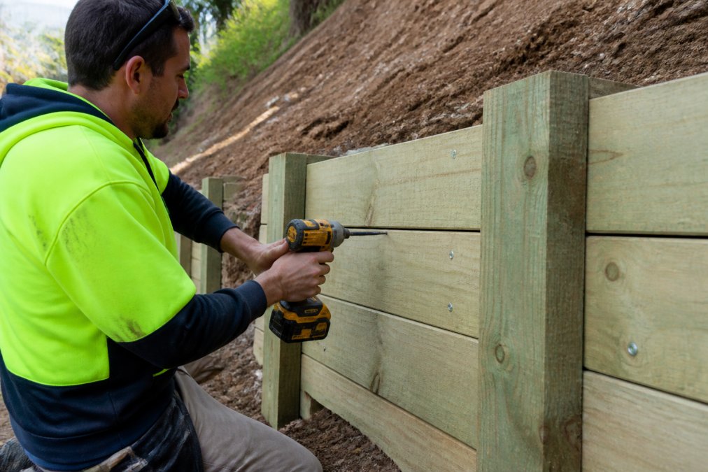 Professional tradesperson using a power drill to install galvanised coach screws through timber sleepers into vertical posts on a sloping site during construction