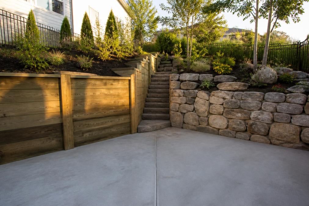 A sloped residential backyard with a pressure-treated timber retaining wall on the left side and a natural stone masonry retaining wall on the right, both bordered by garden beds and a concrete patio, photographed in late afternoon sunlight