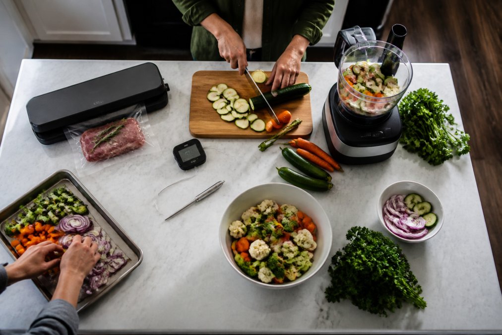 Overhead shot of a busy home kitchen with multiple time-saving gadgets arranged on the counter, smart thermometer, food processor, vacuum sealer, with hands in motion preparing ingredients and vegetables
