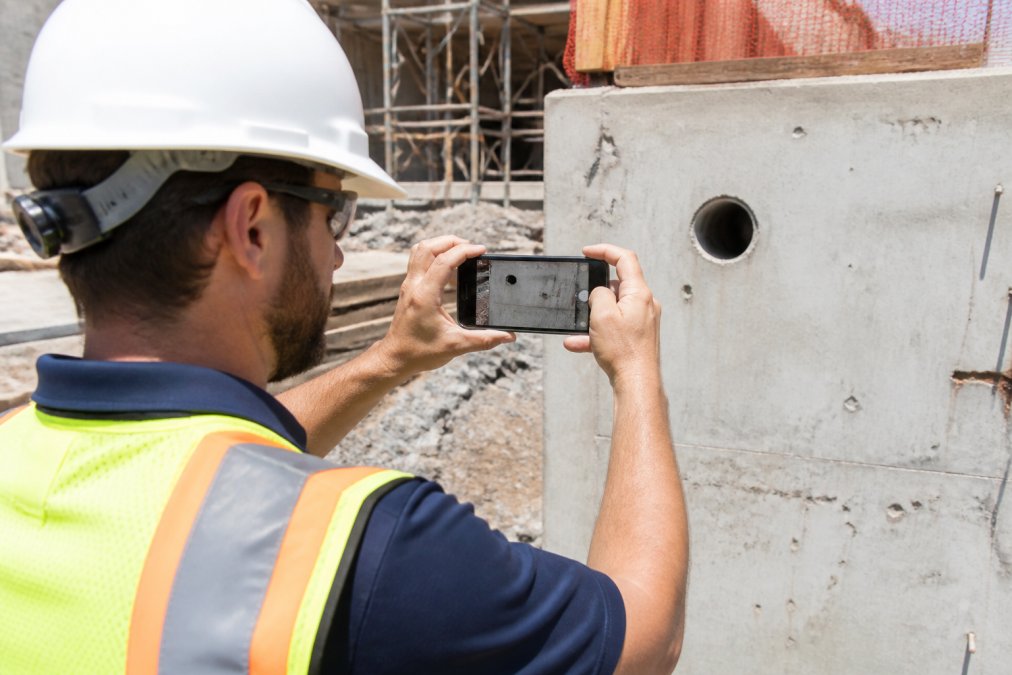 An inspector in a hard hat and hi-vis vest holding an Android smartphone up to photograph a concrete wall detail on a construction site, bright daylight, with the phone screen visible