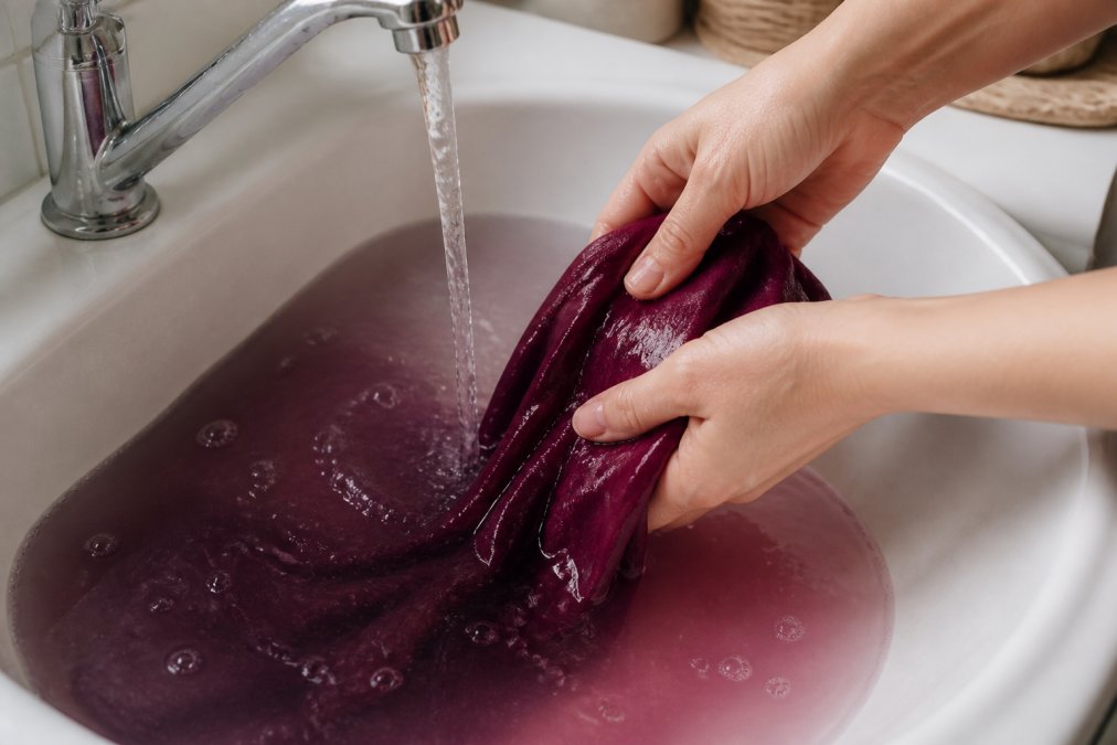 Mani che risciacquano un tessuto appena tinto sotto l'acqua corrente fredda in un lavandino, mostrando il cambio di colore dell'acqua da scuro a trasparente