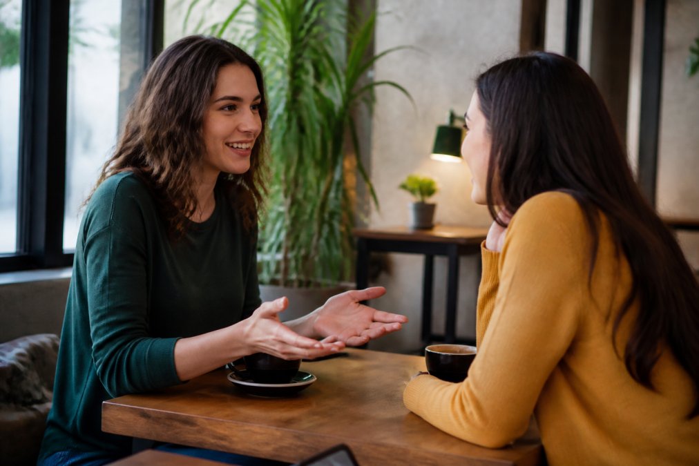 Two friends sitting across from each other at a coffee table, one leaning forward while speaking with an open gesture, both smiling in relaxed conversation suggesting honest, comfortable dialogue