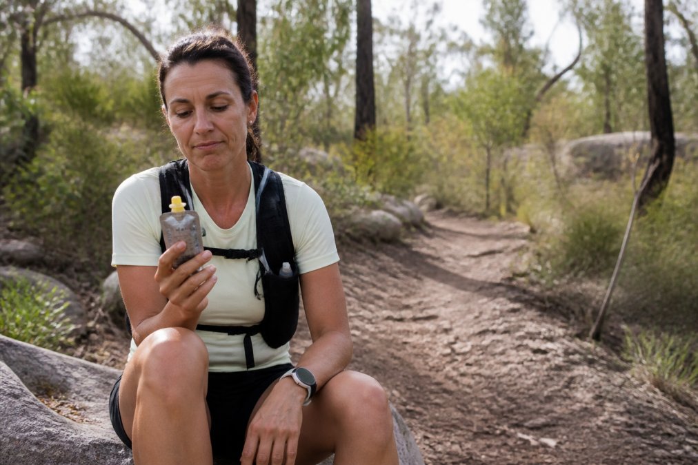 An endurance runner sitting on a trailside rock during a long training run, holding an energy gel packet and looking at it with a tired, unenthusiastic expression, surrounded by native bushland