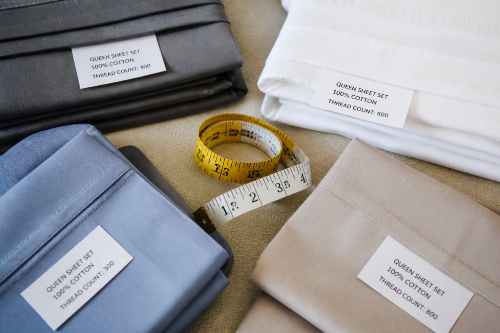Flat lay of folded bedding sets in different colors with a tape measure and product labels visible, showing sizing and thread count details