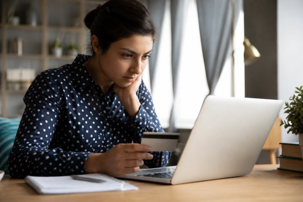 A woman sitting at a desk with a laptop, carefully examining an online clothing store's website, with a credit card and notebook nearby