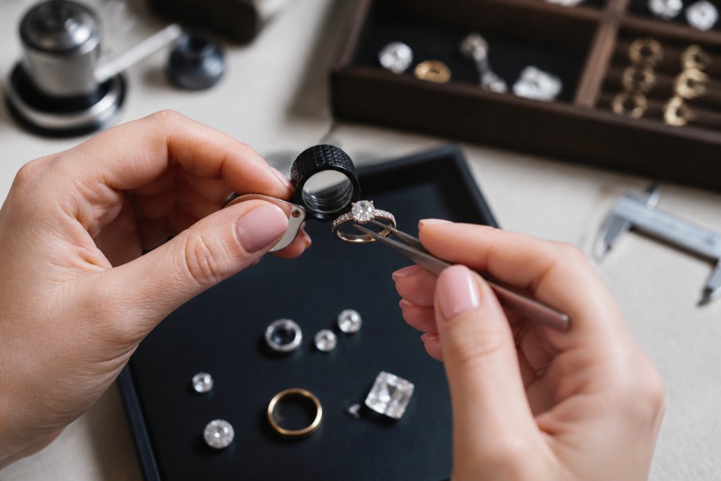 Close-up of hands examining a fine jewelry piece under bright light, showing detailed inspection of hallmarks and gemstone clarity with magnification tools
