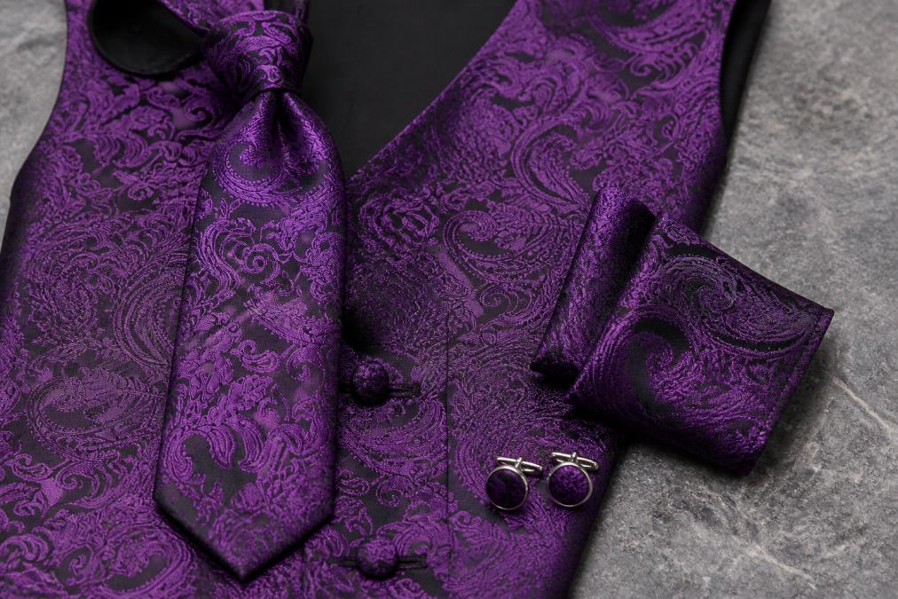Close-up of a groomsman's formal attire showing burgundy waistcoat, coordinated necktie, silk pocket square, and polished cufflinks arranged on a textured surface