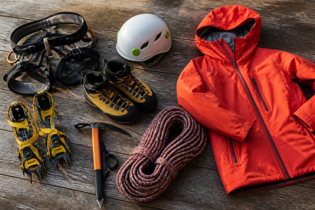 Flat lay of essential mountaineering equipment including harness, helmet, crampons, ice axe, climbing shoes, rope coil, and technical insulated jacket arranged on a weathered wooden surface with natural morning light