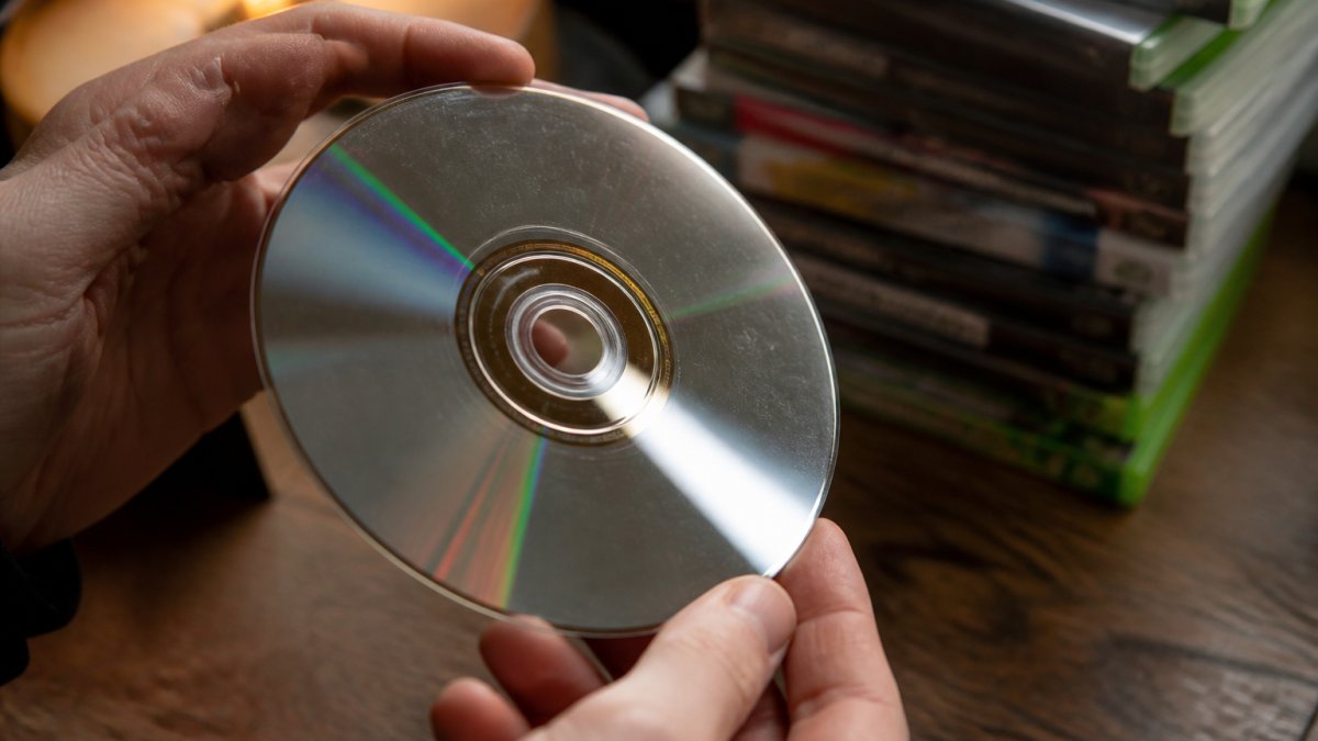 Close-up of hands holding a used game disc up to a light, checking the underside for scratches, with a stack of cartridge cases blurred in the background on a wooden table
