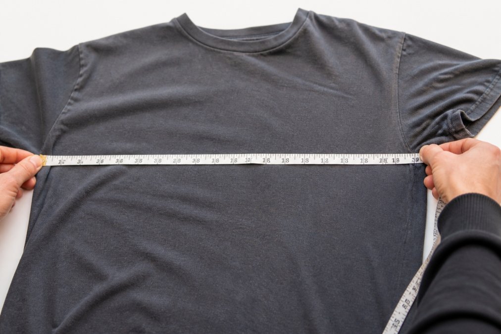 Close-up of hands using a soft measuring tape to measure a vintage t-shirt laid flat on a white surface, showing pit-to-pit measurement across the chest with clear tape markings visible