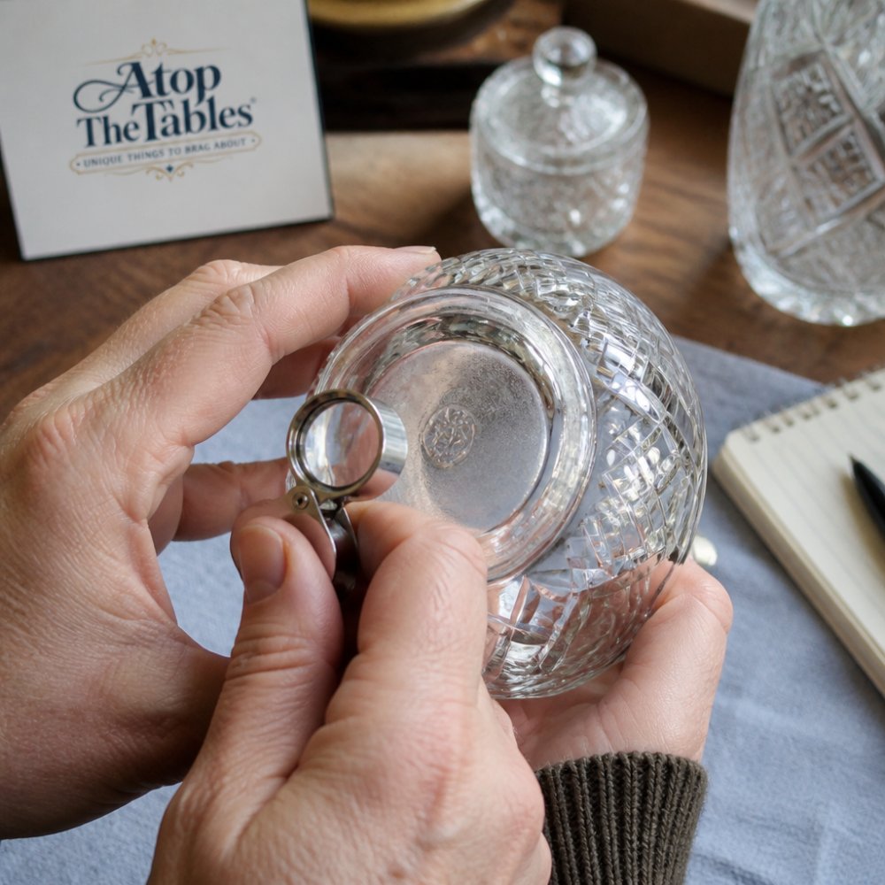Close-up of hands examining a vintage glass piece under natural light, showing inspection of hallmarks and maker's marks on the bottom