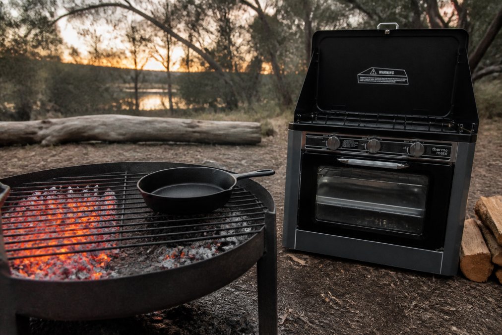 Close-up of a campfire grill grate with glowing charcoal coals arranged in two distinct zones, one side with dense glowing coals for direct heat and one side with sparse coals for indirect heat, a cast iron skillet resting on the indirect side in a bush camping setting at dusk