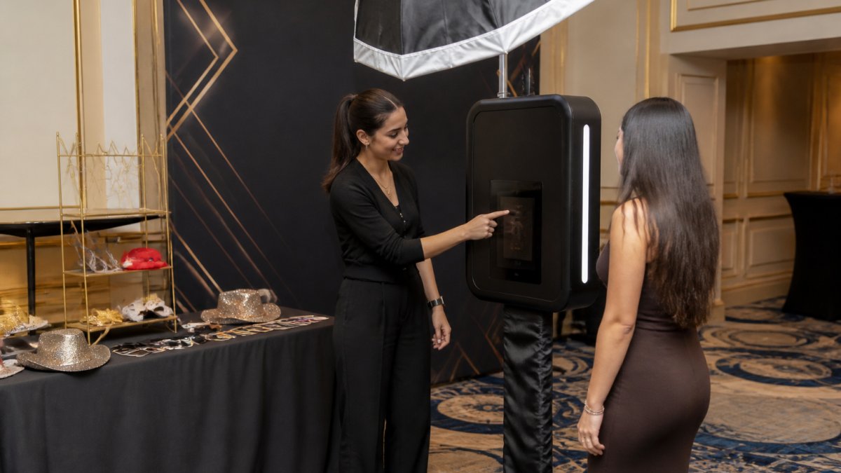 Professional photo booth setup in elegant event space with custom branded backdrop featuring company logo and event colors, custom props displayed on table, attendant in professional attire assisting guest at tablet interface with studio lighting overhead