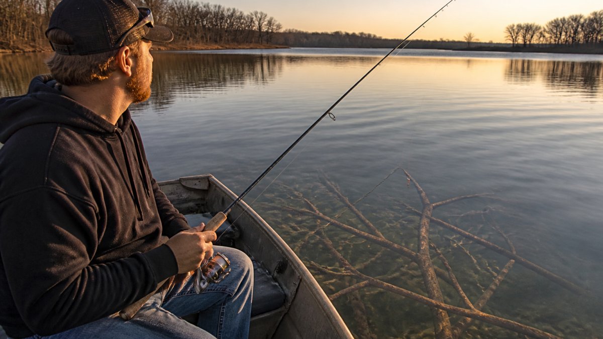 An angler in a small aluminum boat on a calm lake at golden hour, holding a light spinning rod with a slip bobber rig, submerged brush visible just below the surface beside the boat
