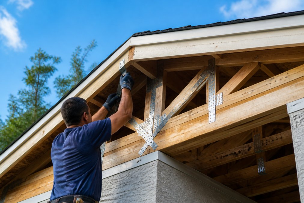 Contractor installing metal hurricane straps and reinforced roof trusses on residential home exterior, with close-up view of roof attachment hardware and structural reinforcements on a clear day