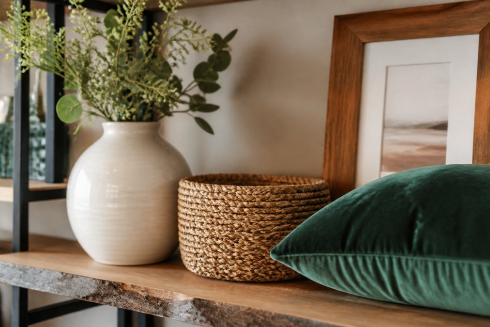 Close-up of a styled shelf displaying layered textures: smooth ceramic vase in white, woven natural fiber basket, soft velvet cushion in jewel tone, and wooden picture frame with warm finish, demonstrating how different tactile materials create visual interest in one cohesive arrangement