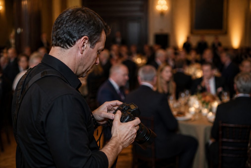 Professional photographer adjusting camera settings at a dimly lit formal dinner event, checking aperture and ISO on the camera body while guests converse in soft background lighting