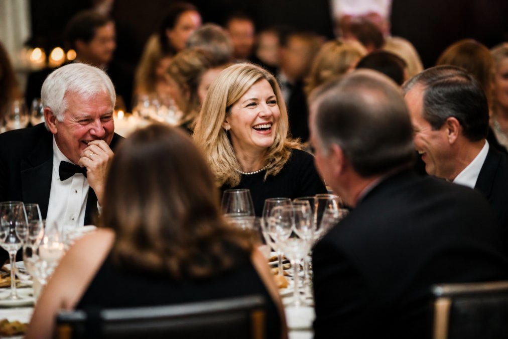 A candid moment of donors laughing and interacting naturally at a gala table, captured from a discreet distance with soft available light, showing genuine emotion and connection