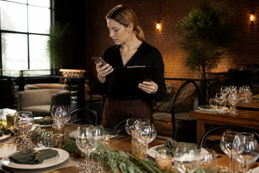 Event coordinator or host checking clipboard and mobile phone while arranging table settings and decor in a private event space with careful attention to detail