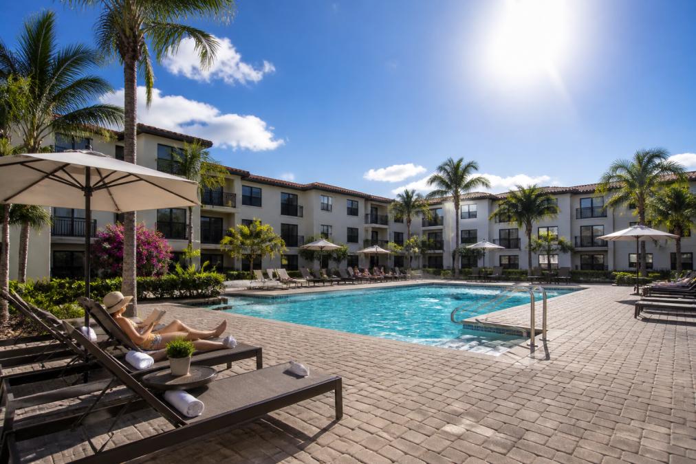 A bright, sunny Florida afternoon with blue skies over a modern apartment complex featuring a resort-style pool and residents enjoying outdoor spaces, lounge chairs, and landscaped gardens under intense sunshine