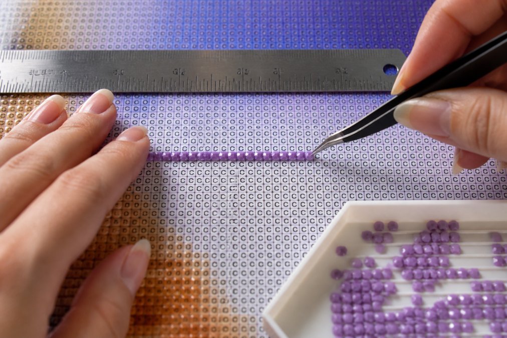 Close-up of hands positioning diamond drills in a straight horizontal line on a canvas, using tweezers with a metal ruler visible as a guide in the background, natural lighting showing the grid pattern clearly