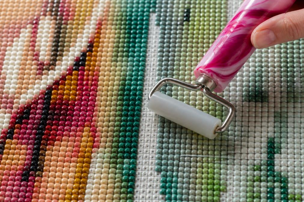 Overhead view of a diamond painting canvas showing a section with small gaps between drills, with a roller tool positioned to compress and close the spacing, demonstrating the technique
