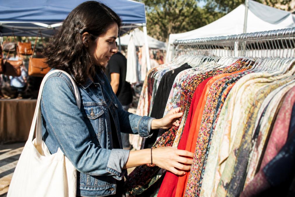 A woman smiling as she browses through a rack of colorful vintage and secondhand clothing at an outdoor thrift market on a bright sunny day, warm natural light illuminating the garments