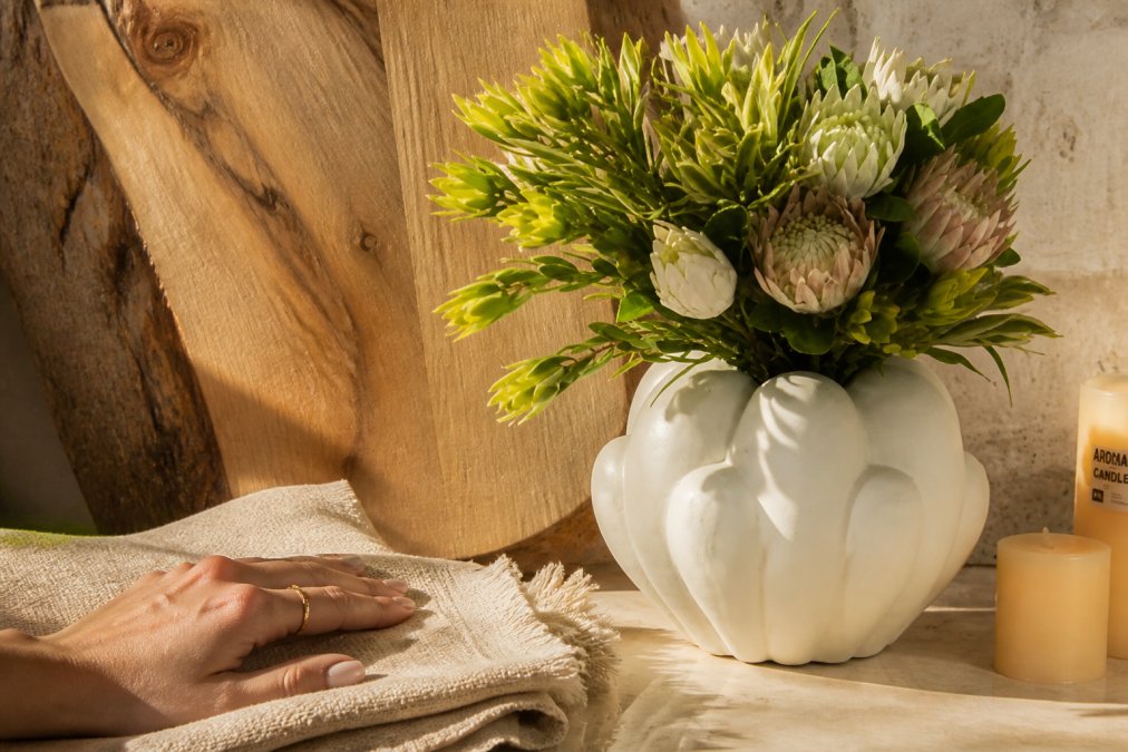 Close-up of a hand touching layered natural textures, linen fabric with visible weave, warm wood grain showing natural variations, and smooth stone surface, arranged on a neutral surface with warm afternoon lighting