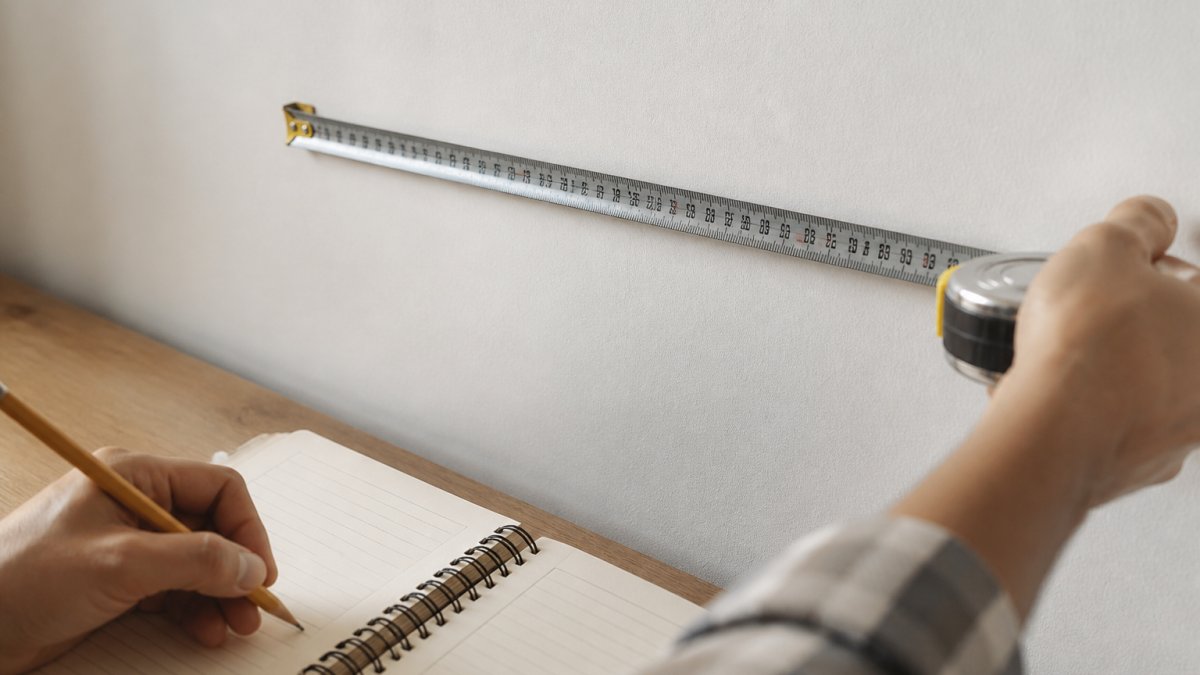 Close-up of a metal measuring tape stretched across a white wall with a notebook and pencil recording dimensions on a wooden surface nearby
