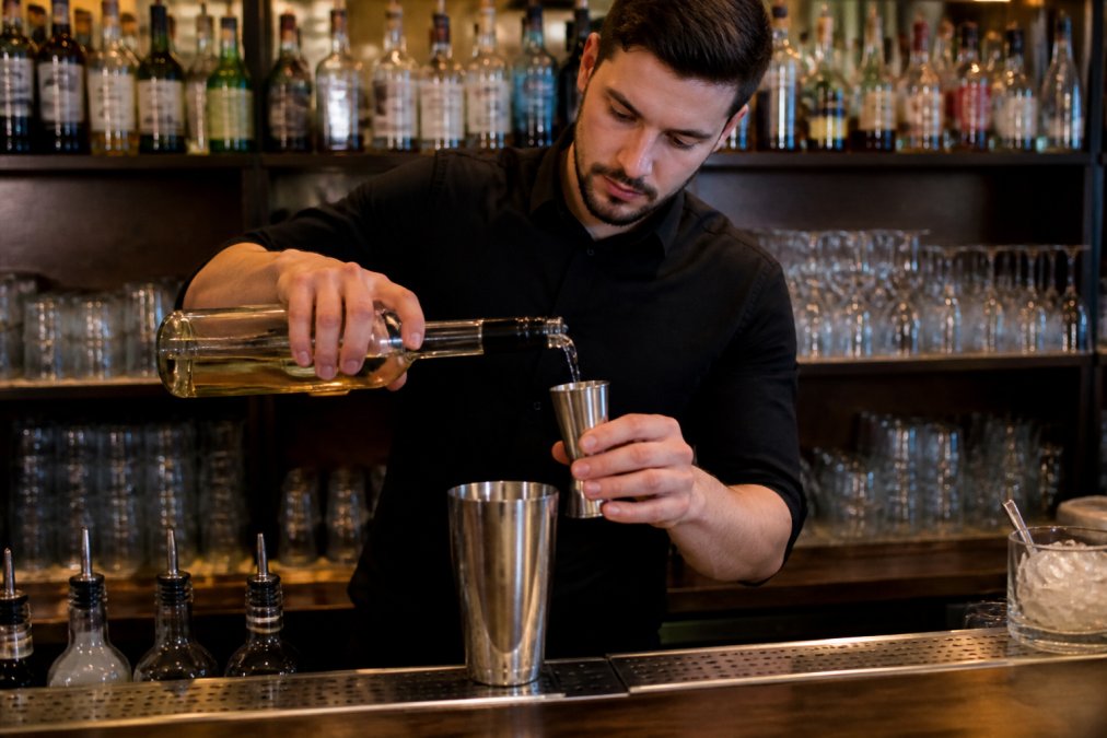A bartender carefully measuring a spirit pour using a stainless steel jigger at a busy bar counter, with shelves of bottles and glassware visible in the background