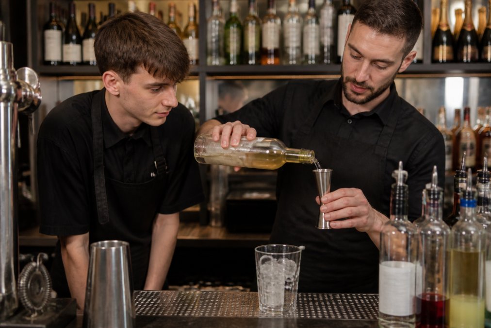 A bartender demonstrating proper jigger technique to a newer team member at a well-lit bar counter, with bottles and training materials visible