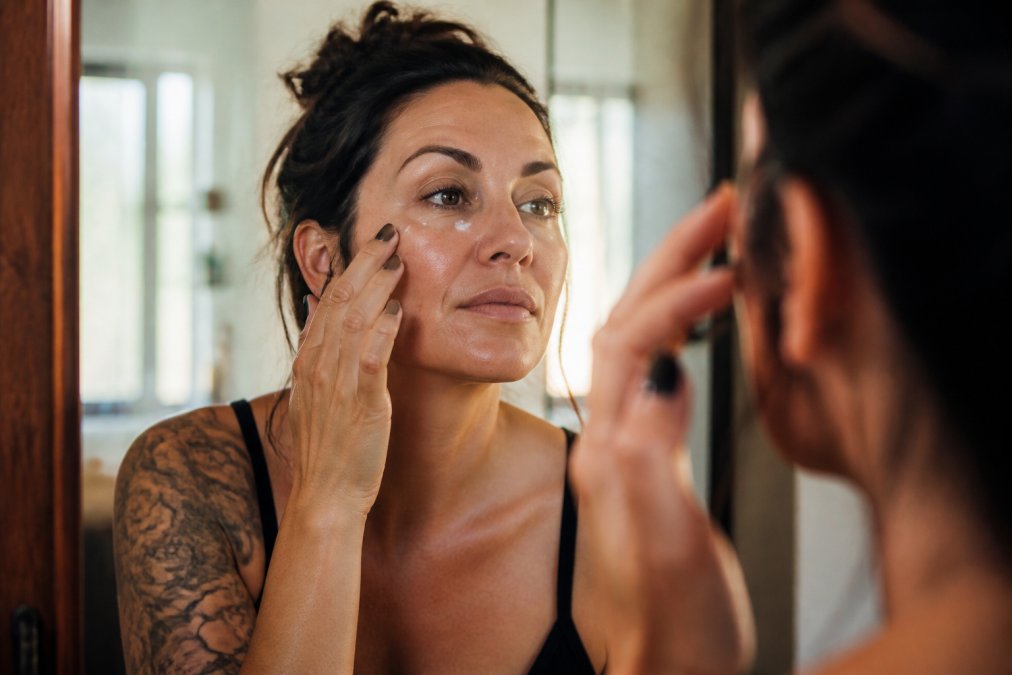 Woman in her 40s applying under-eye cream in soft morning light, looking at herself in mirror with a relaxed, confident expression