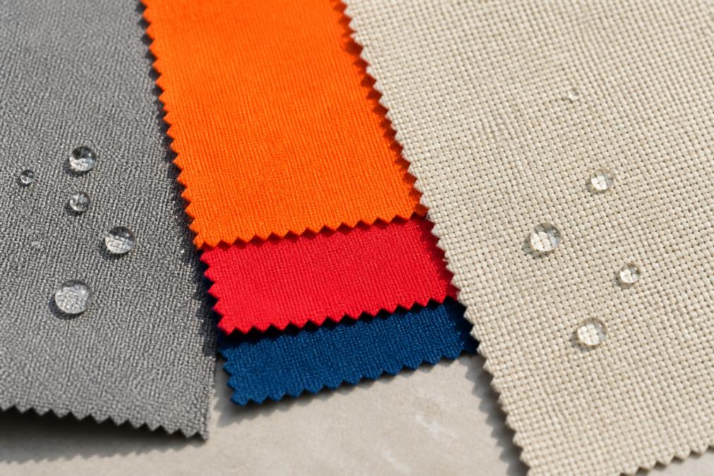 Close-up of fabric swatches showing texture differences between matte polyester with water beading on surface, bright Sunbrella acrylique in vibrant colors, and natural-looking olefin fibers with water droplets, displayed in natural daylight