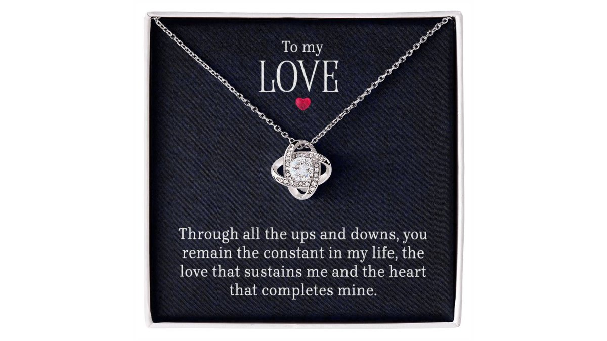 A woman's hands gently holding a delicate gold love knot necklace pendant, soft natural light, warm and intimate atmosphere