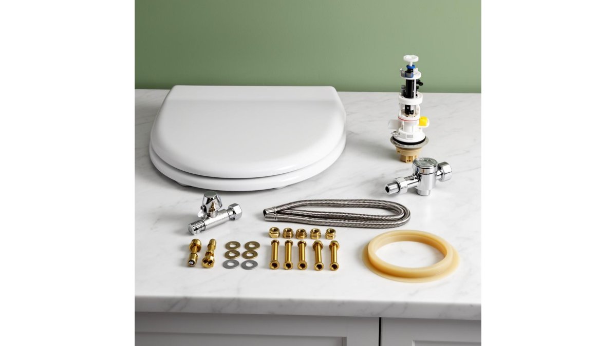 A maintenance technician in a professional uniform installing a toilet fill valve inside a commercial restroom, with a pre-kitted parts box open on the floor beside him