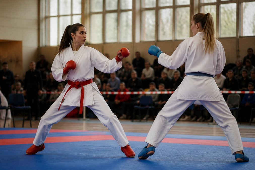 Professional karate athlete in kumite stance wearing lightweight gi, mid-movement during sparring match on tatami mat with natural gym lighting