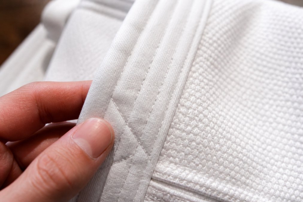 Close-up of karate uniform fabric showing dense canvas weave texture and reinforced stitching detail, with fingers touching the material to demonstrate premium quality and construction