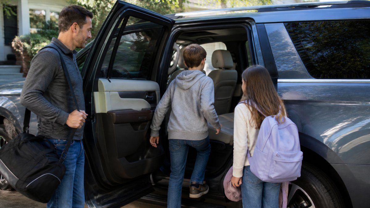 A family with two children loading into a spacious three-row SUV through an open rear door, second-row bench seat visible, bright daytime driveway setting