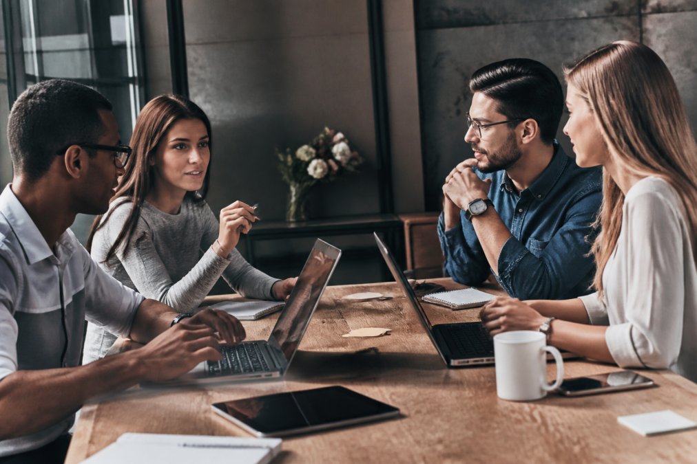 Marketing team collaborating around a table with laptops and tablets, reviewing campaign strategy and analytics dashboards displayed on monitors