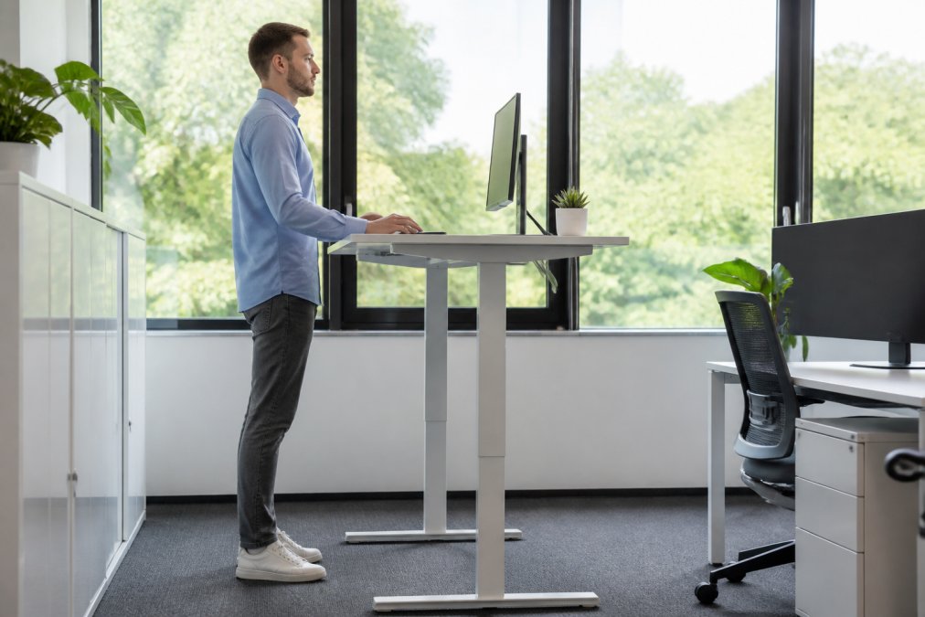 Person working at ergonomic standing desk with proper posture, natural office lighting from window, monitor at eye level, keyboard and mouse at elbow height