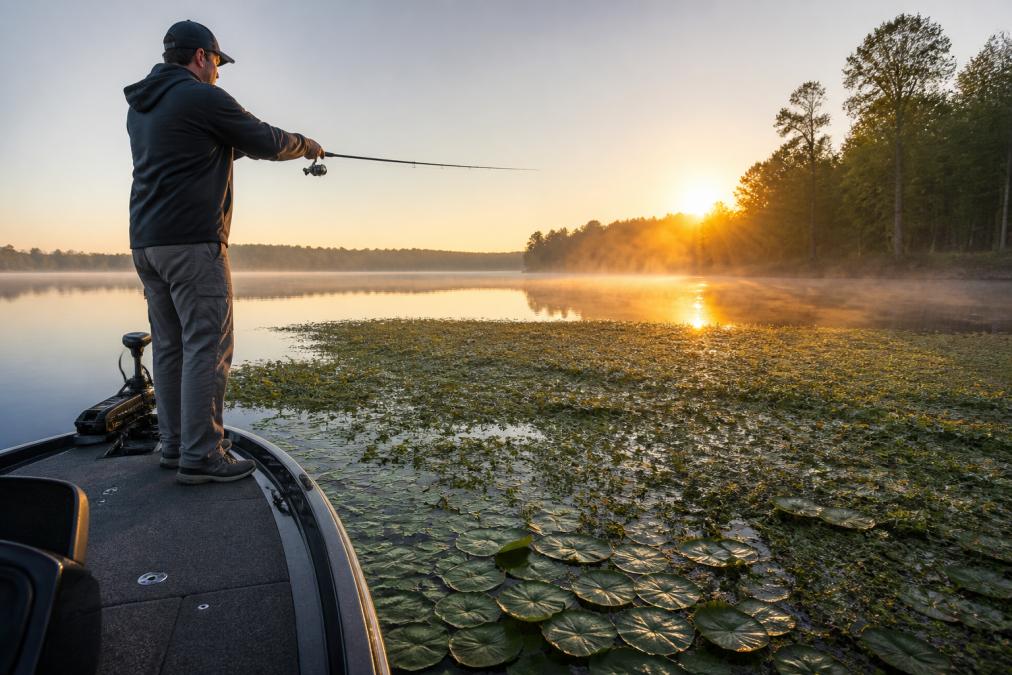 Angler casting weedless lures into thick matted vegetation at sunrise on a calm lake with lily pads