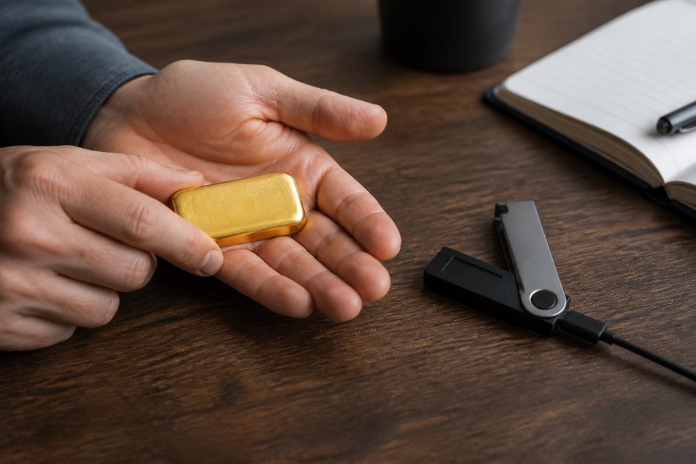 Hands holding a small gold bar next to a hardware wallet for gold and bitcoin storage on a desk
