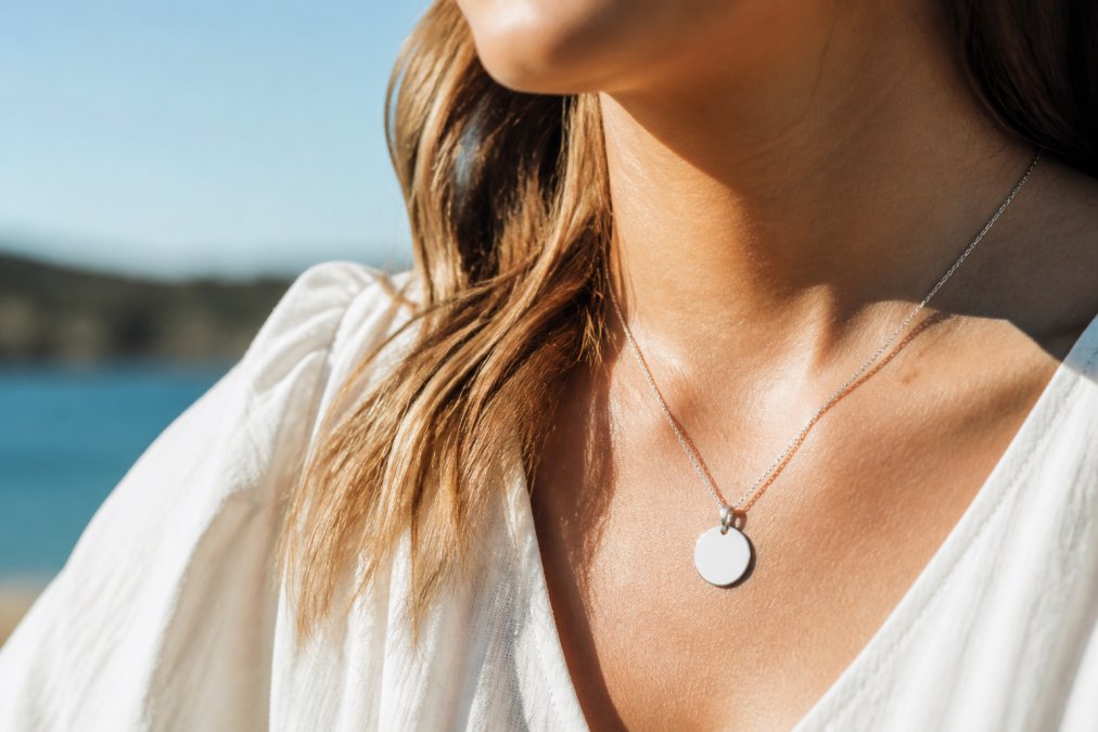 Woman wearing a polished stainless steel pendant necklace with custom engraving, the pendant catching natural sunlight against her collarbone, photographed in bright outdoor setting