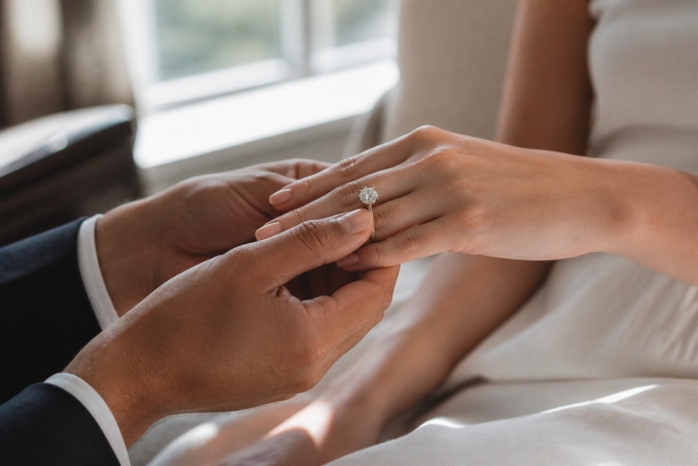 A close-up of a couple's hands, with the man gently placing an engagement ring on the woman's finger. Soft, natural window light, elegant and timeless atmosphere. Focus on the ring's sparkle.