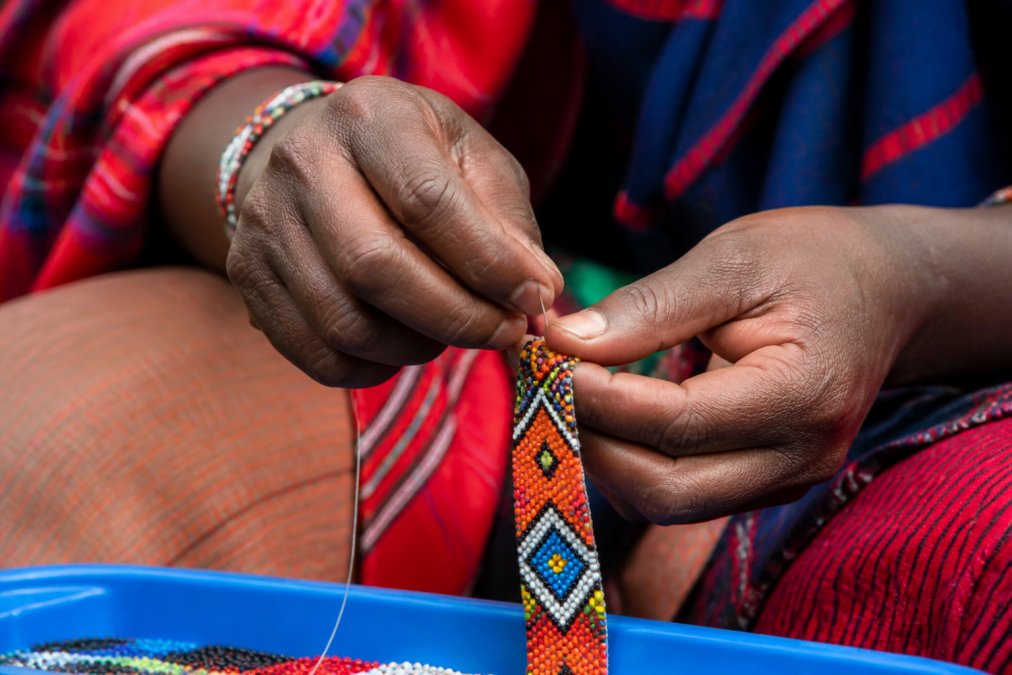 Close-up of artisan hands carefully threading colorful beads onto wire, showing intricate geometric patterns with traditional textile colors visible in the background, natural daylight