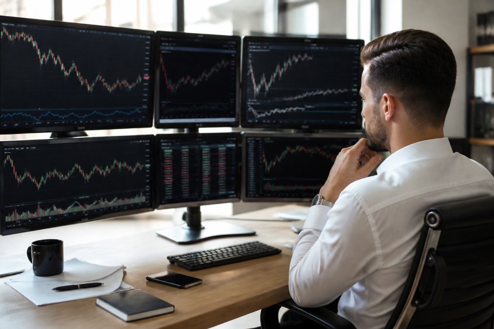 Professional trader at desk with multiple monitors displaying real-time forex charts, technical indicators, and currency pair price movements in bright office setting