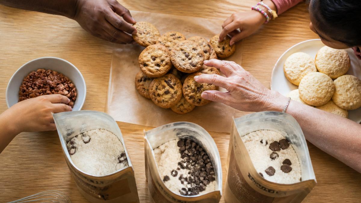 Overhead flat-lay of a light-wood kitchen counter showing three open gourmet cookie mix pouches, a stack of baked chocolate chip cookies on parchment, a small bowl of pecan halves, and a plate of pale teacakes, with a diverse set of hands of different ages and skin tones reaching in from the edges