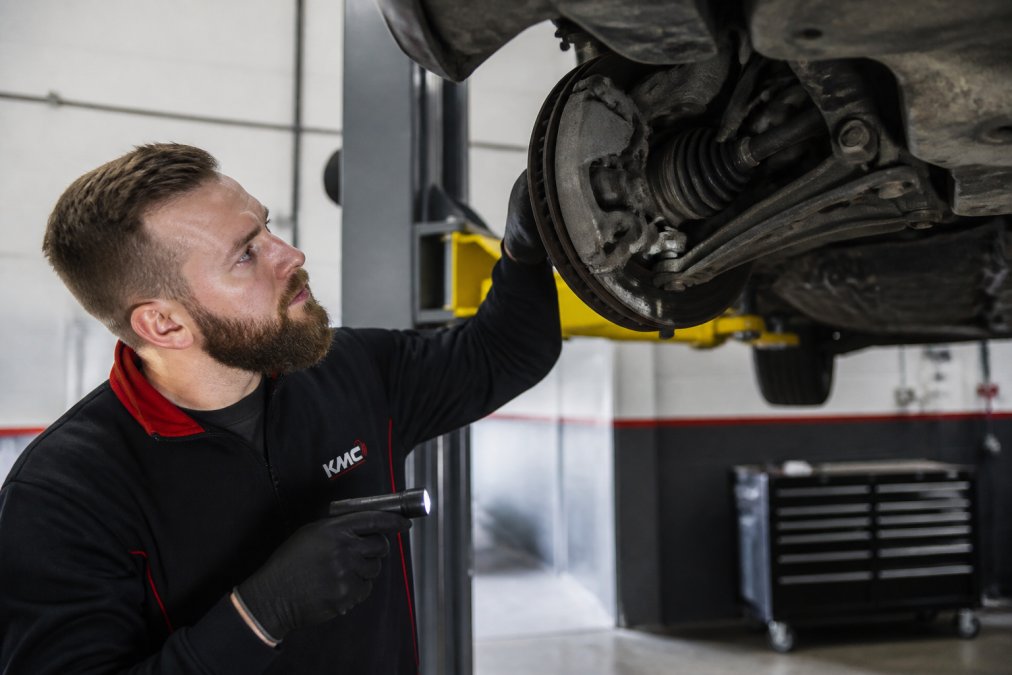 Mechanic inspecting a vehicle's brake components and suspension during an MOT test, with diagnostic equipment visible in the background