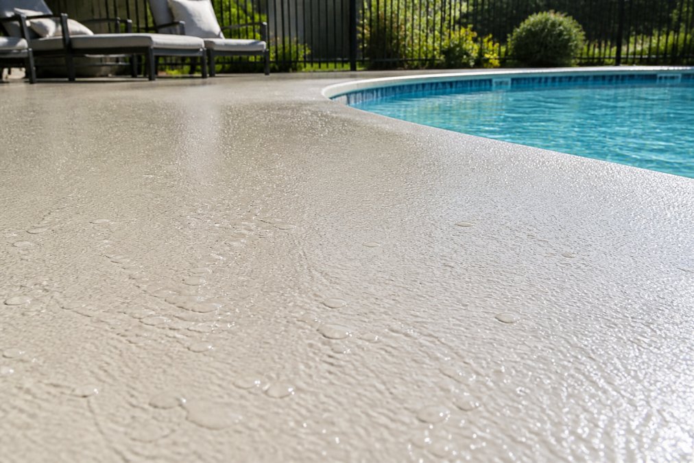 Residential pool deck with fresh polyaspartic cool-touch coating showing slip-resistant textured finish in bright afternoon sunlight, water droplets visible on the surface demonstrating water resistance, pool edge and lounge chairs in background