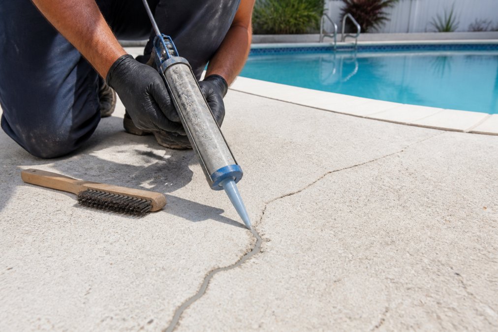 Technician applying concrete crack repair compound into visible hairline crack on pool deck surface, showing surface preparation with wire brush and application of flexible sealant material